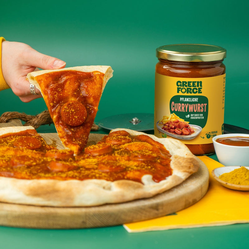 Person nimmt sich ein Pizzastück getoppt mit veganem Currywurst Readymeal von Greenforce, Packung steht daneben