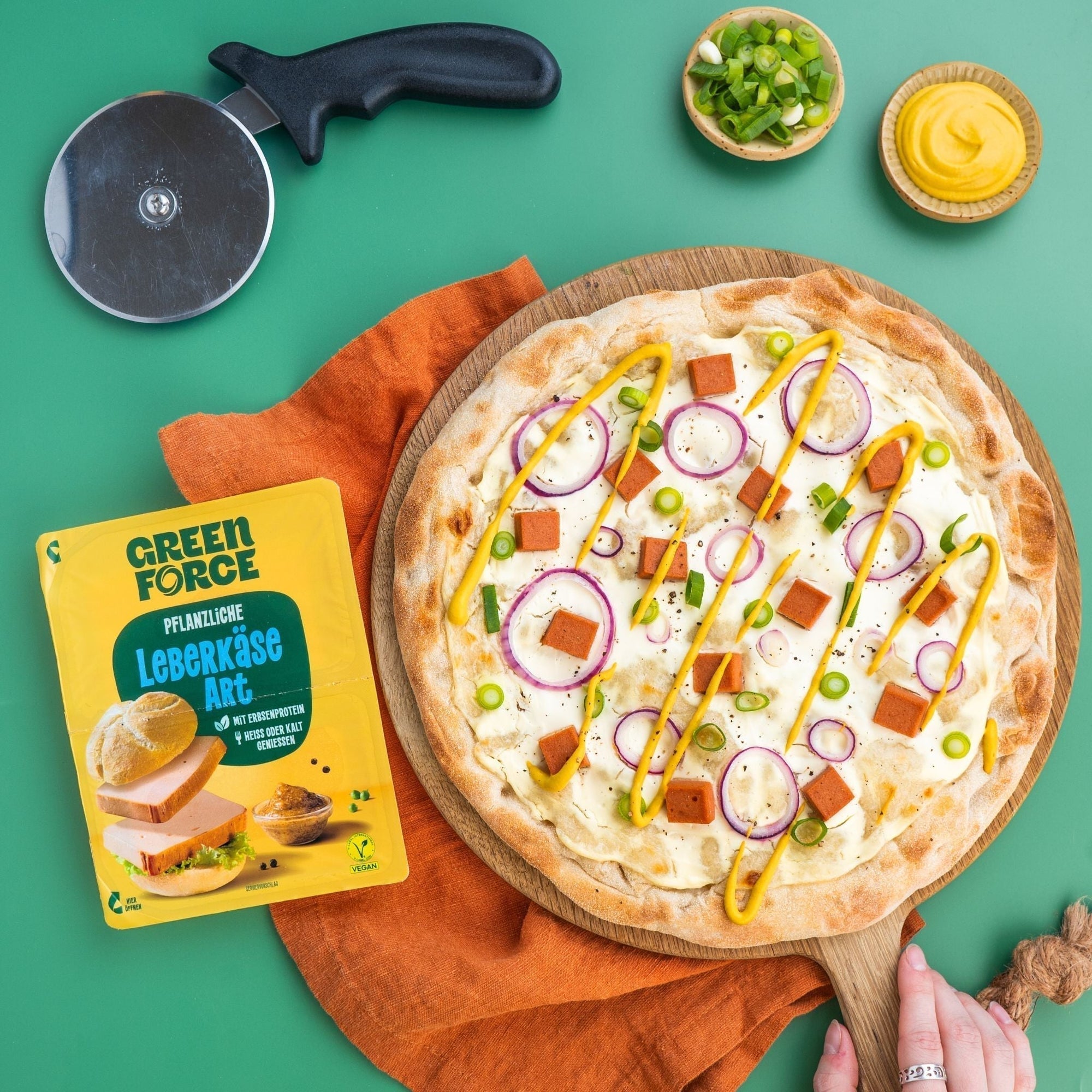 Vegane Leberkäse Pizza getoppt mit veganem Creme fraiche, Zwiebelringen, veganem Leberkäse, Frühlingszwiebeln und Senf. Greenforce Leberkäse Packung daneben.