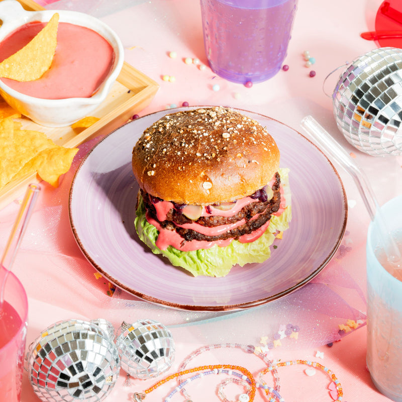 Belegter veganer Burger mit Nachos, pinker Käsesauce und Getränken im Hintergrund