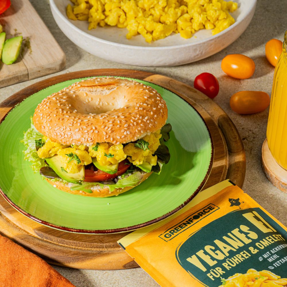 Breakfast Bagel mit veganem Ei, Avocado, Tomate und Salat auf Teller mit weiteren Zutaten und der veganen Greenforce Ei Packung um den Teller herum.