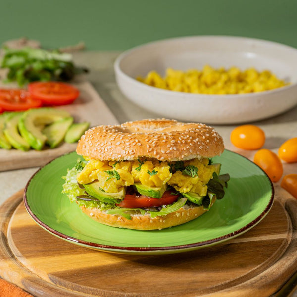 Veganer Breakfast Bagel mit veganem Ei, Avocado, Tomate und Salat mit weiteren Zutaten um den Bagel herum.