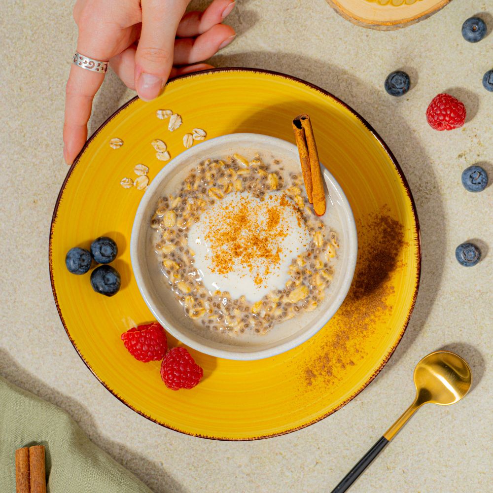 Overnight Oats mit veganem Haferdrink serviert in Schüssel mit veganem Joghurt, Zimt und Beeren. Person hält Teller mit Servierschale und Toppings.