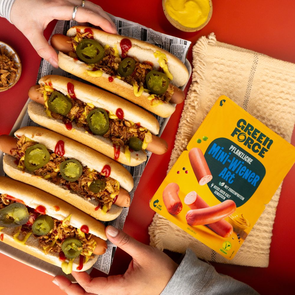 Zwei Hände greifen sich veganen Hot Dog mit veganen Wienern, Ketchup, Senf, Röstzwiebeln und Jalapenos, daneben liegt die Greenforce Mini Wiener Packung