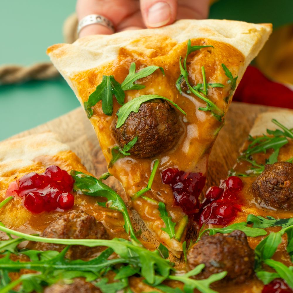 Close Up Pizzastück getoppt mit pflanzlichen Köttbullar, Cremesauce, Wildpreiselbeeren und Rucola.