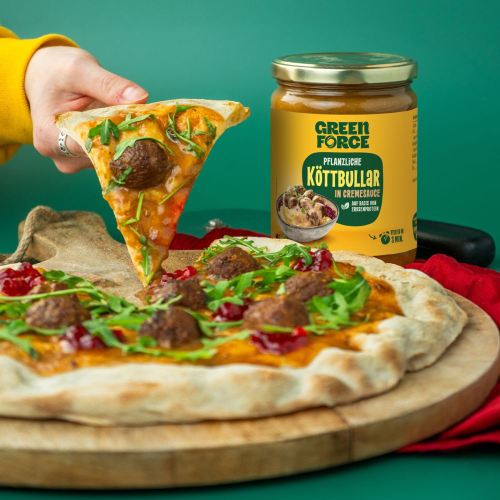 Person nimmt sich ein Stück von einer Pizza getoppt mit pflanzlichen Köttbullar, Cremesauce, Wildpreiselbeeren und Rucola. Greenforce Köttbullar Packung steht dahinter.