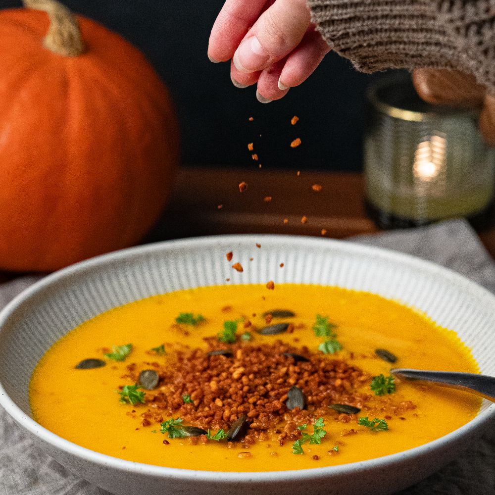 Hand streut vegane Bacon Bits auf Kürbissuppe