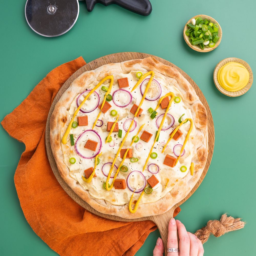 Vegane Leberkäse Pizza getoppt mit veganem Creme fraiche, Zwiebelringen, veganem Leberkäse, Frühlingszwiebeln und Senf.