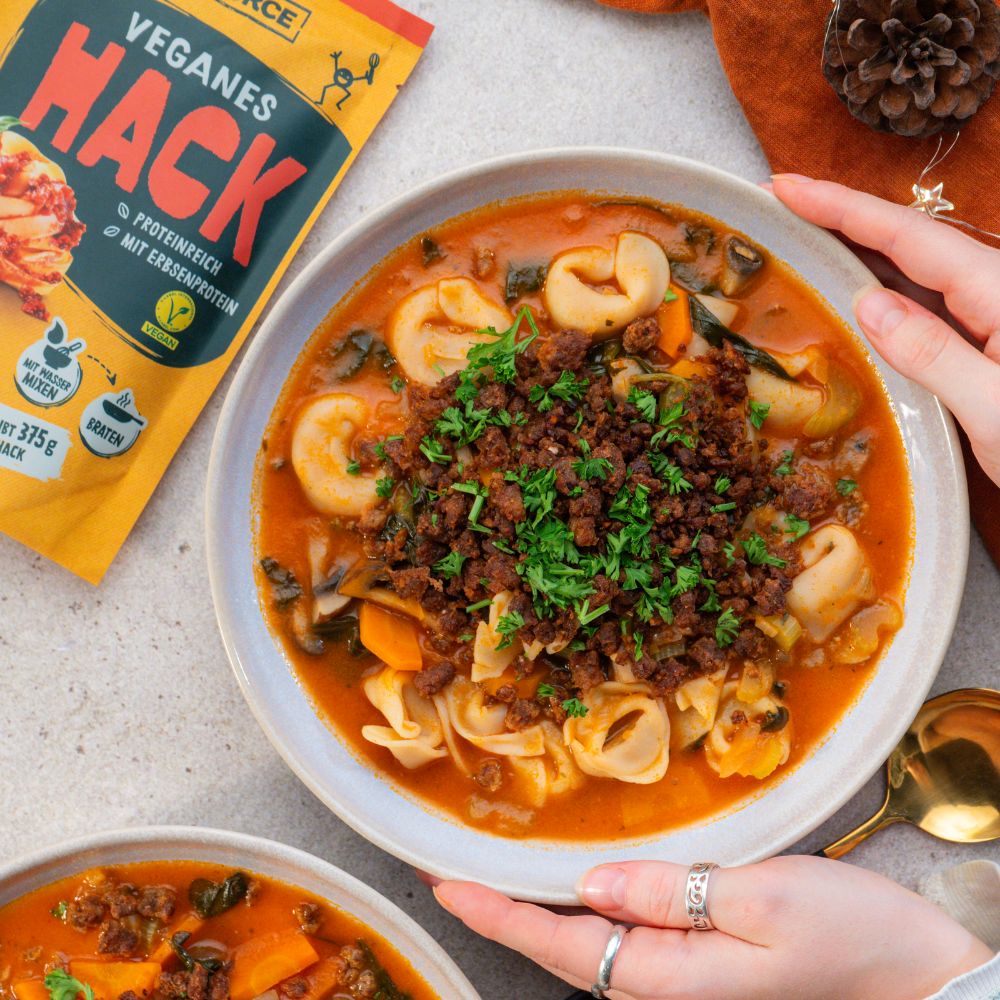 Hände umgreifen Teller mit Tortellini Suppe getoppt mit veganem Hack und Petersilie, daneben liegt eine Greenforce Hack Packung