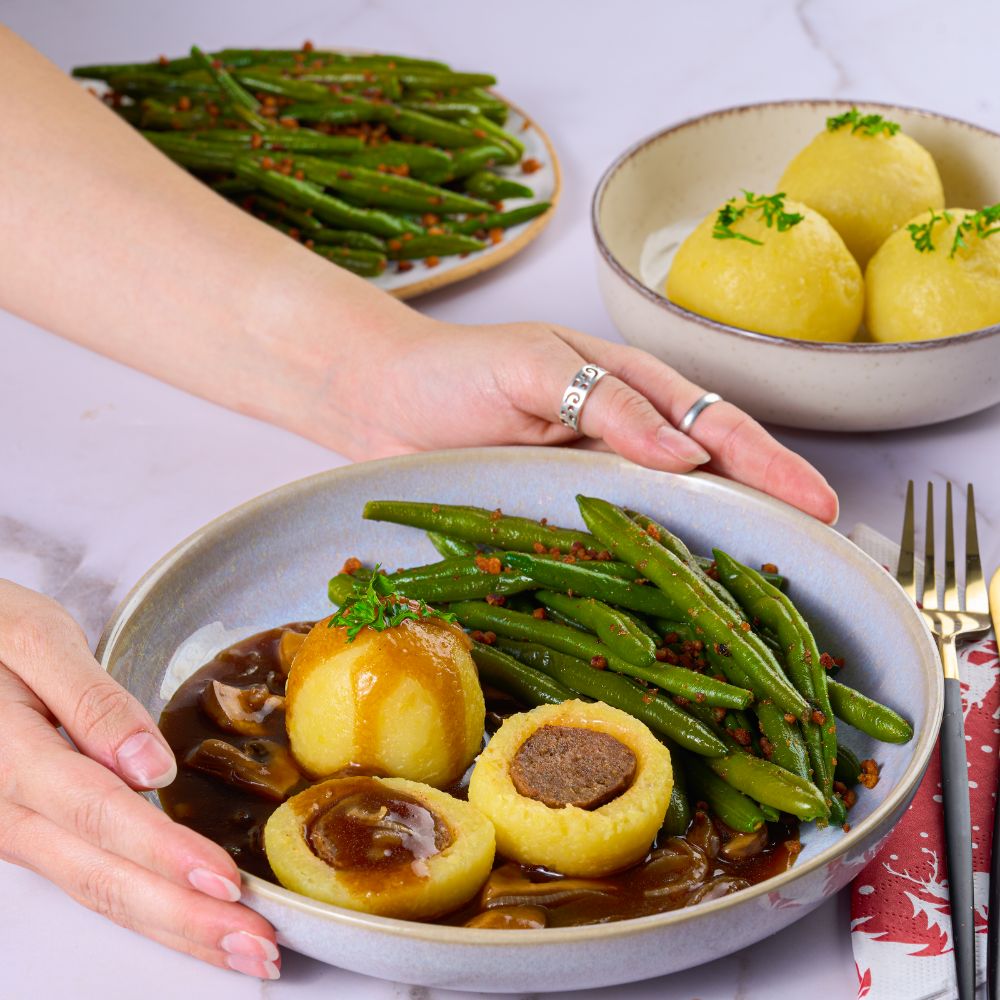 Hände umschließen Teller mit veganem Weihnachtsfestessen mit Knödeln, Bratensoße und Speck Bohnen