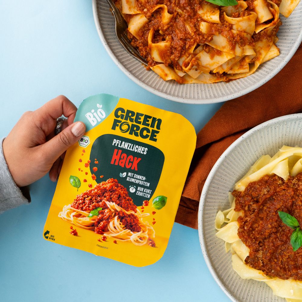 Zwei angeschnittene Teller mit Pasta mit veganer Bolognese, daneben hält eine Hand die Greenforce Hack Packung